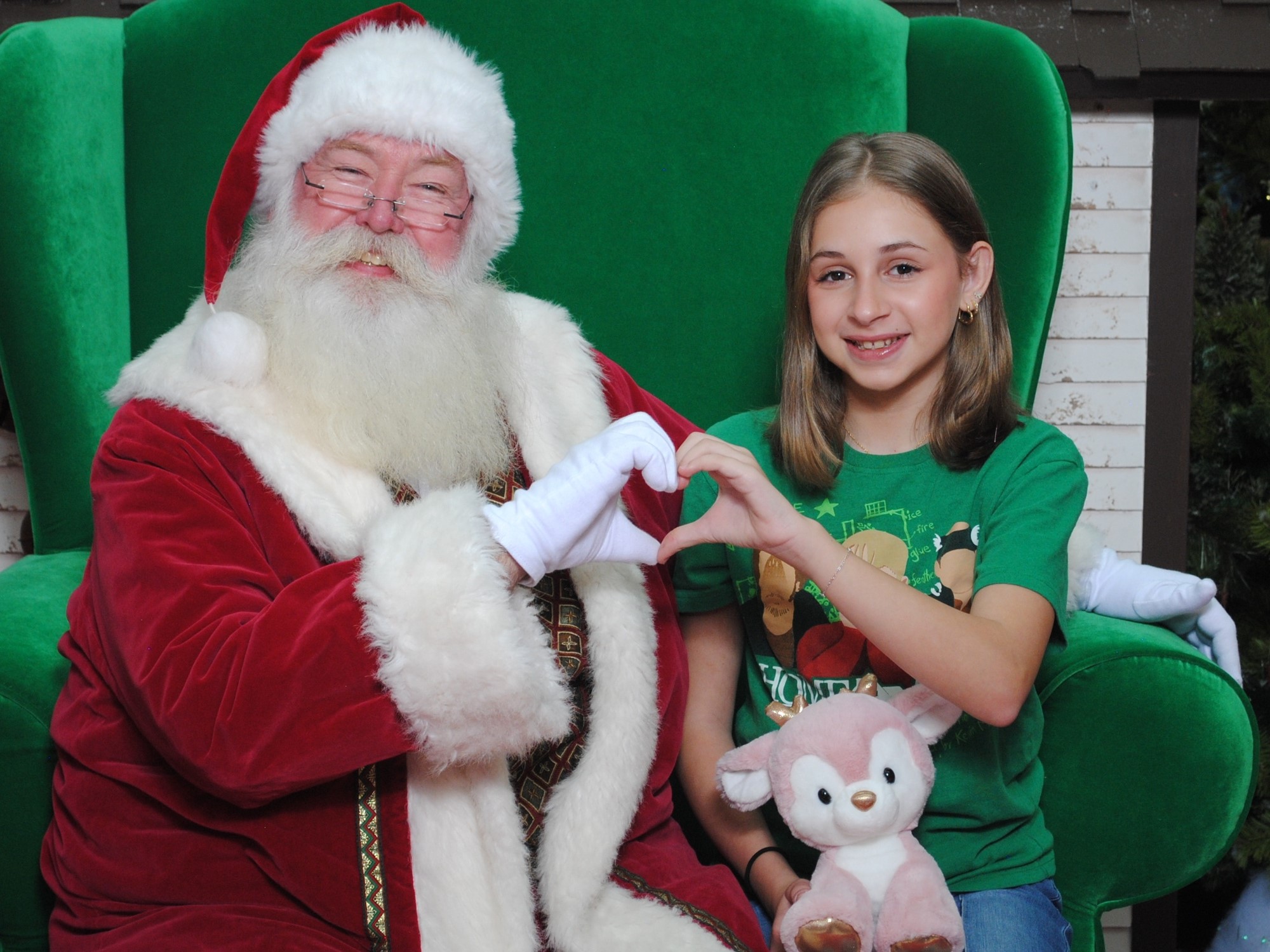 Santa Claus and a child create a heart shape with their hands, sitting together on a green chair, surrounded by festive decorations.